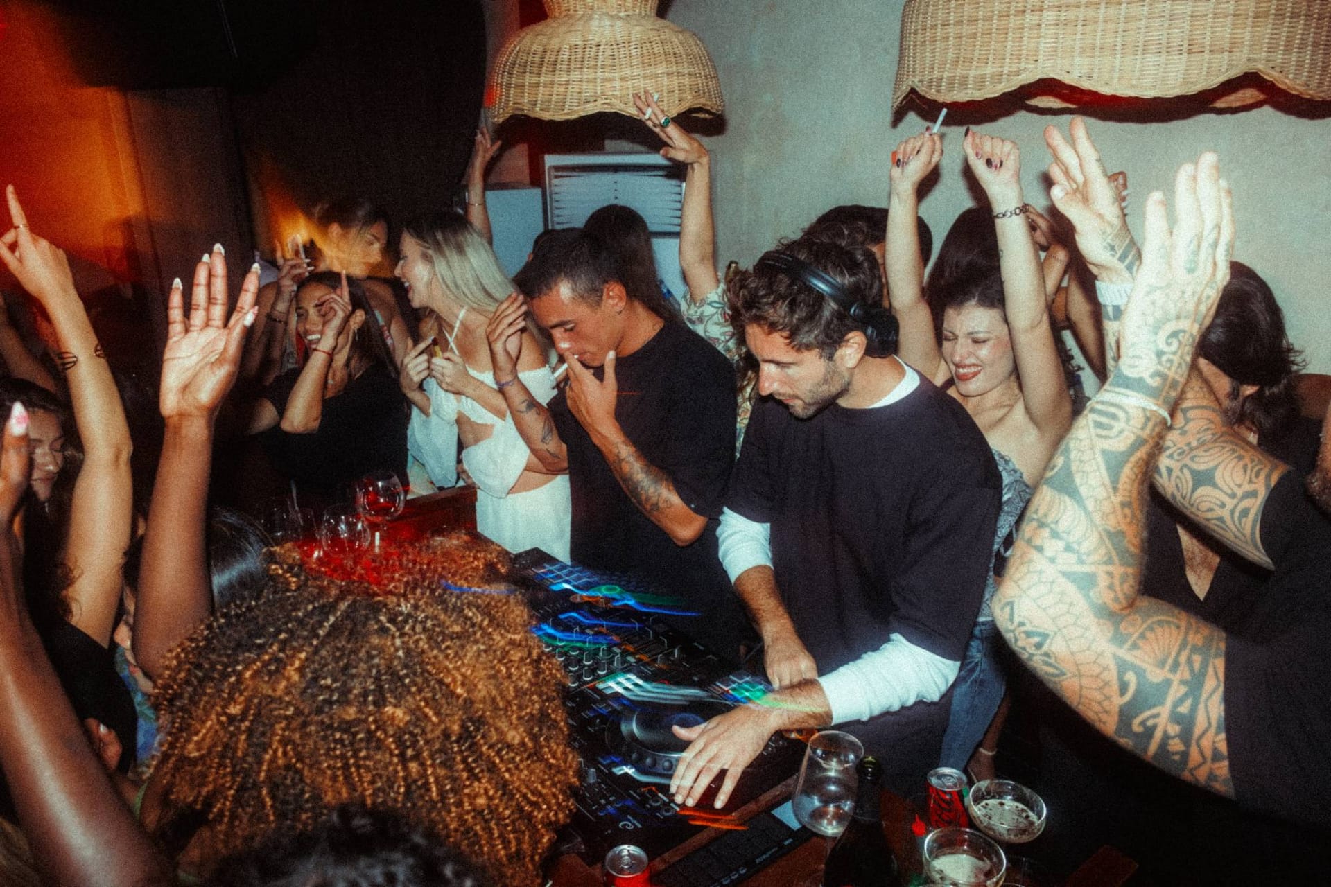 Two DJs at the decks, guests dancing with their hands in the air under the rattan lamps